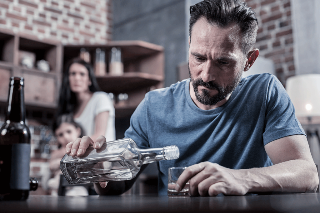 Person with alcohol use disorder pouring drink in front of family causing trauma from alcoholic parents