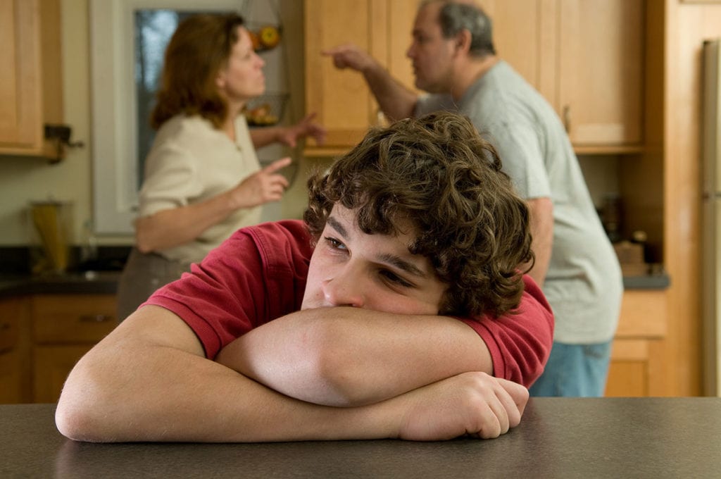 a young man sits at a kitchen counter as his parents fight in the background and thinks about teen depression rates among children of divorce