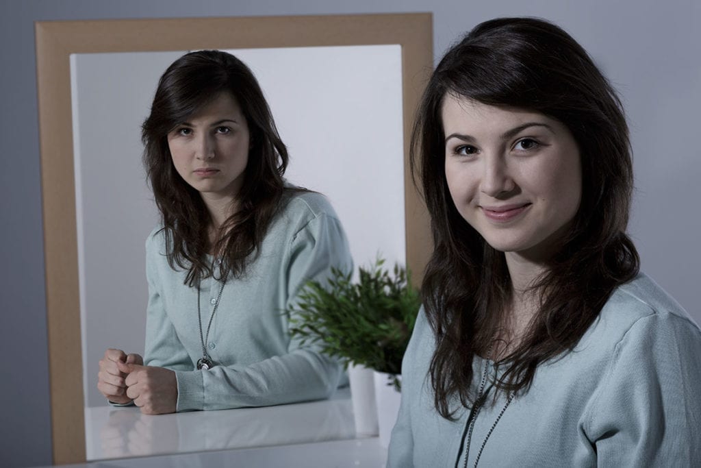 a woman wonders whats the difference between bipolar 1 and bipolar 2 as she stands in front of a mirror
