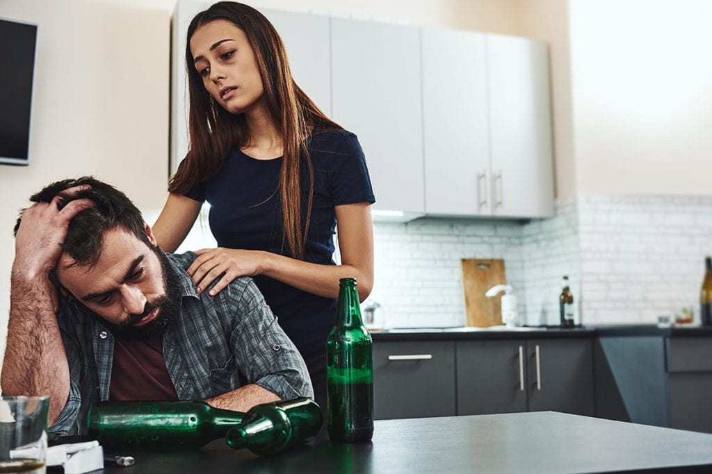 woman comforts man in kitchen surrounded by empty bottles while questioning if it's physical vs psychological dependence