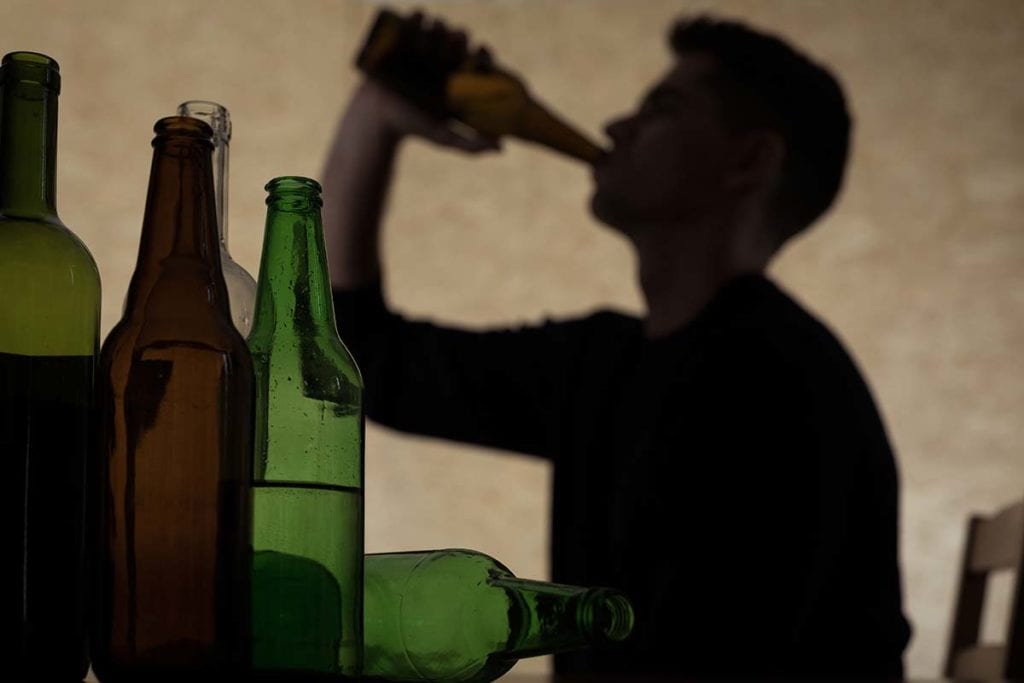 A large number of bottles are in the forefront, while in the background a man drinks, and wonders how addictive is alcohol