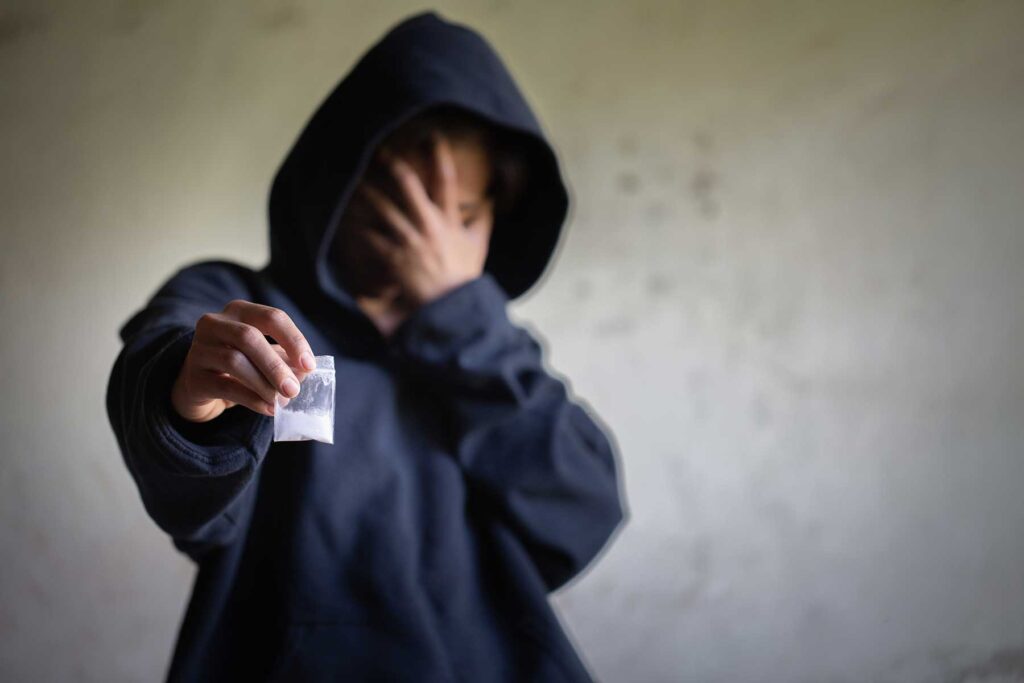 Person wearing hoodie, holding a bag of cocaine, and covering face with hand, wondering, "Why do some people get addicted to cocaine, while others don't?"